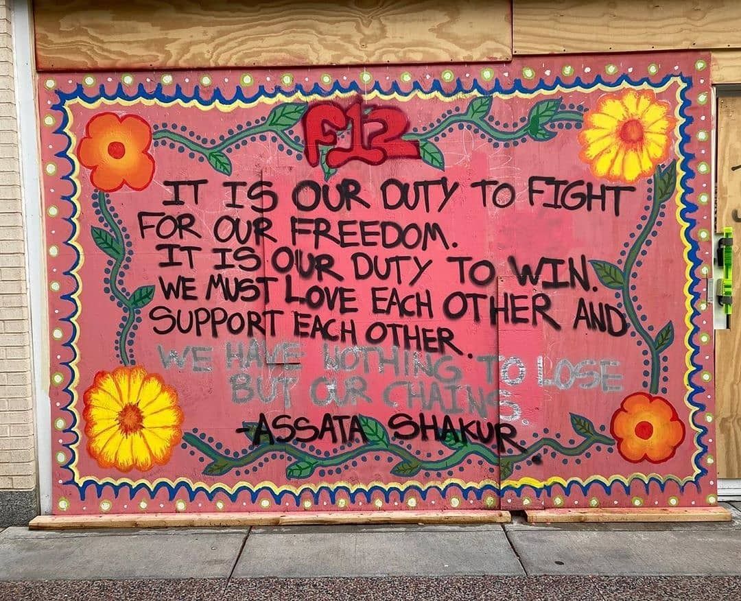 A large piece of street art that is salmon pink with flowers around the outside and a large "F12" at the top. In black script it says: It is our duty to fight for our freedom. It is our duty to win. We must love each other and support each other. We have nothing to lose but our chains. - Assata Shakur"